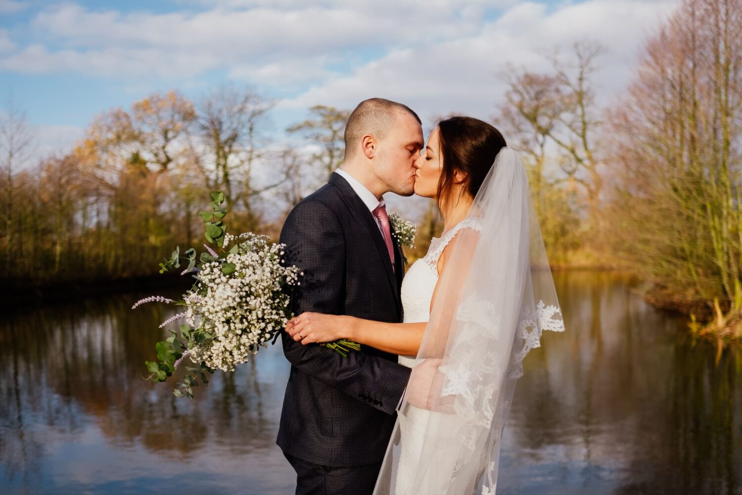bride and groom kiss at styal lodge lake lake in sun