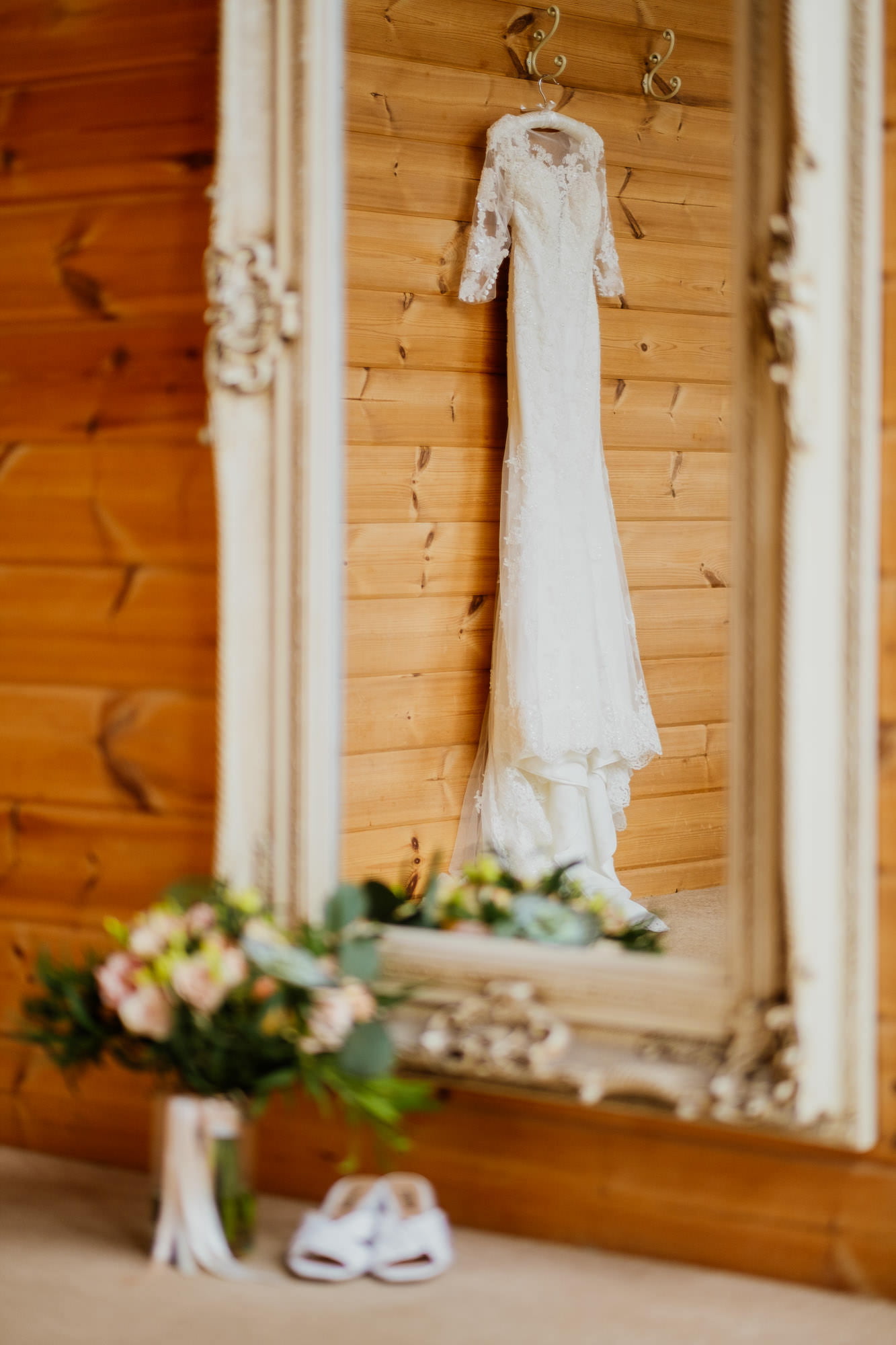 wedding dress hanging in full length mirror with shoes and flowers