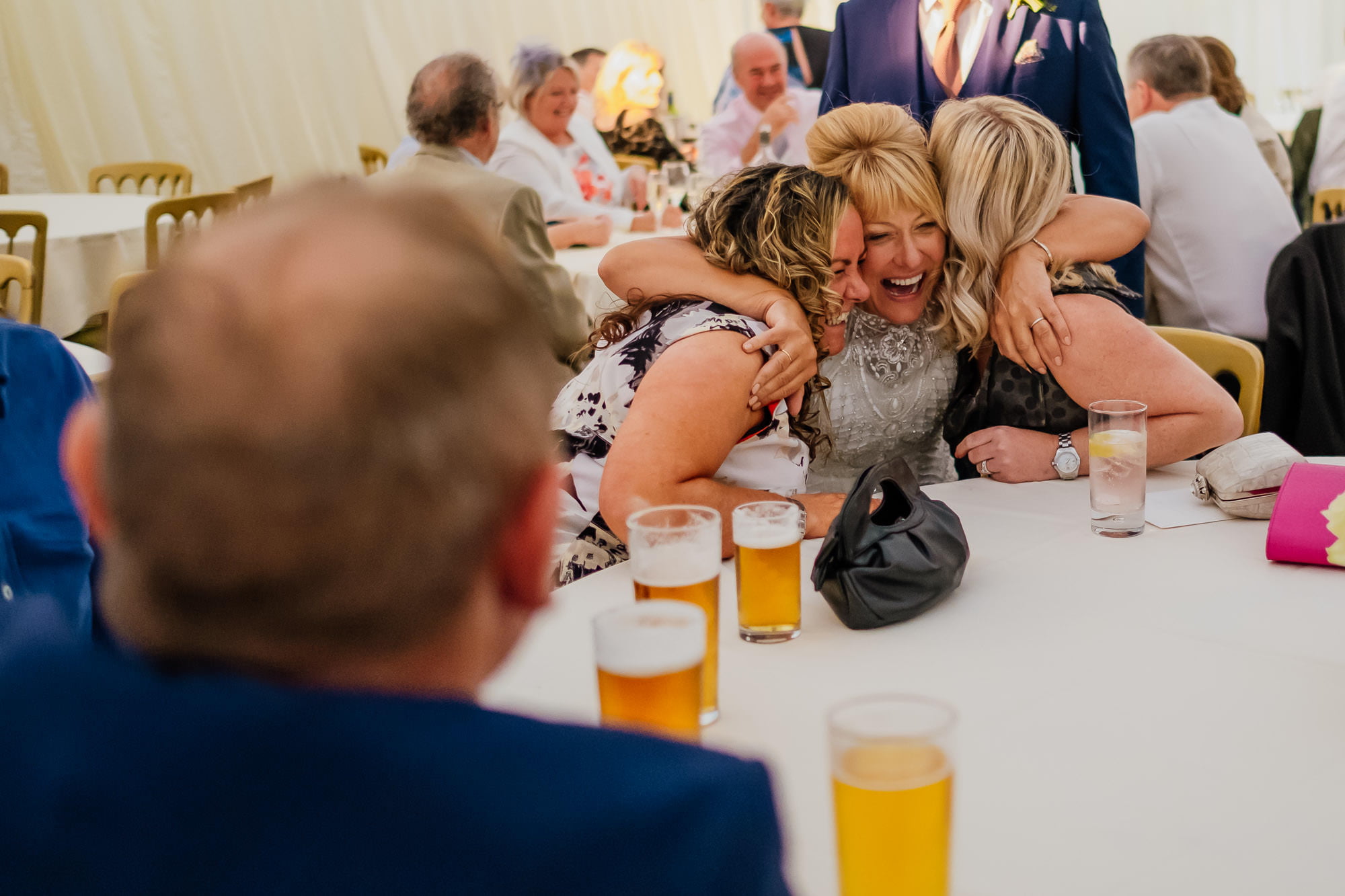 Bride hugging two wedding guests sat at a table