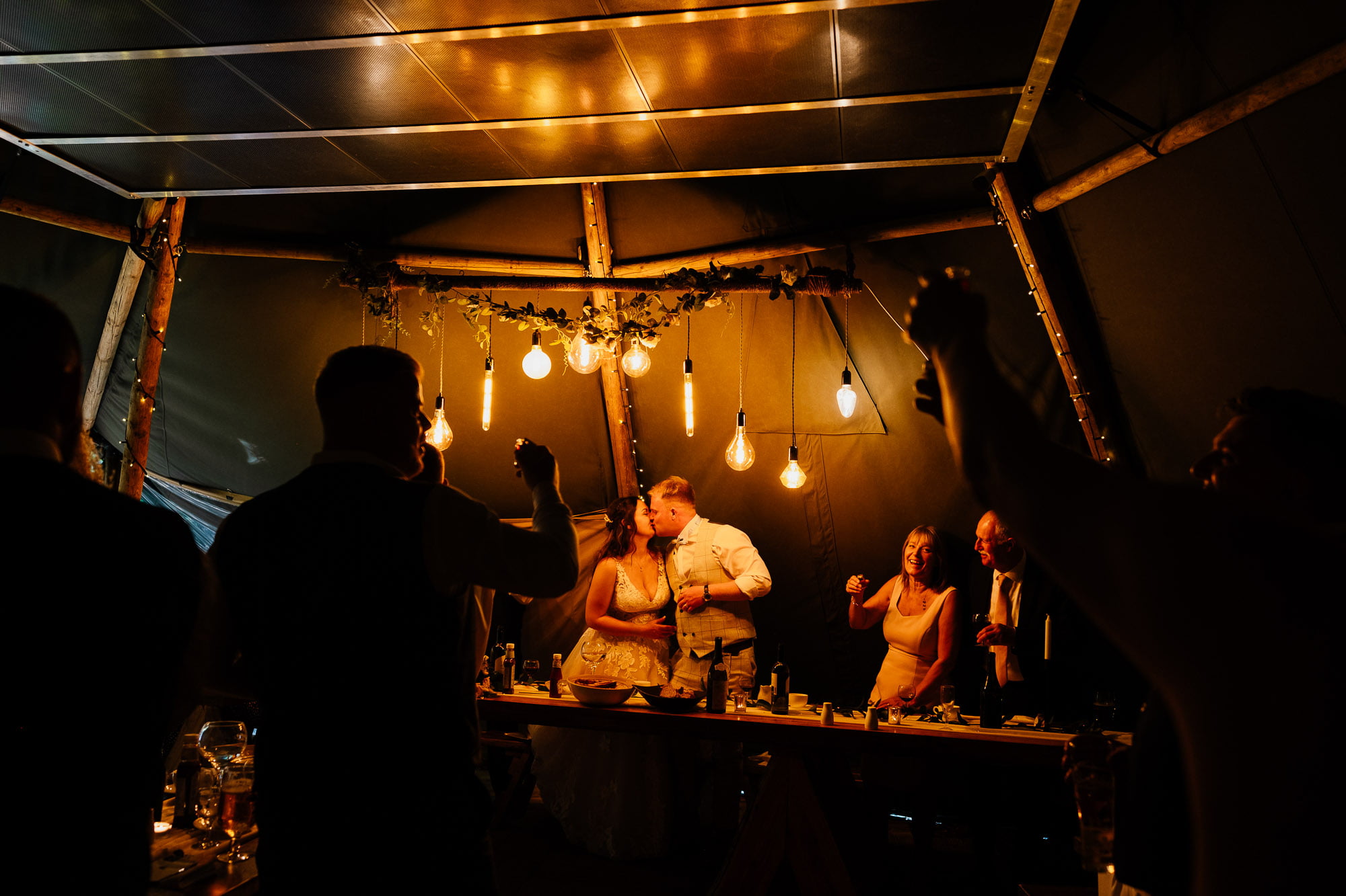 Bride and groom kiss under edison lights in wedding tipi