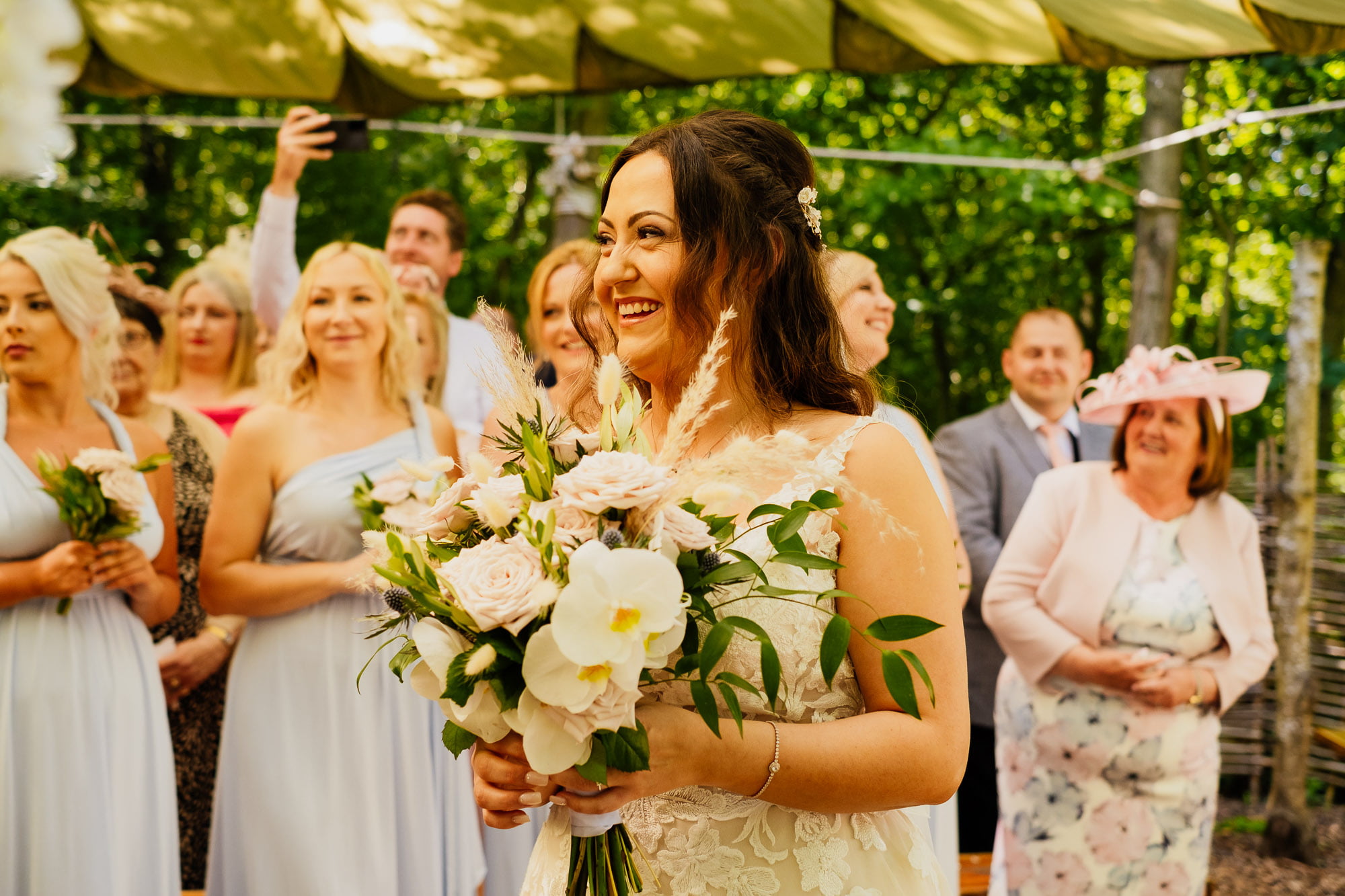 bride smiling holding flowers with guests in the background during ceremony