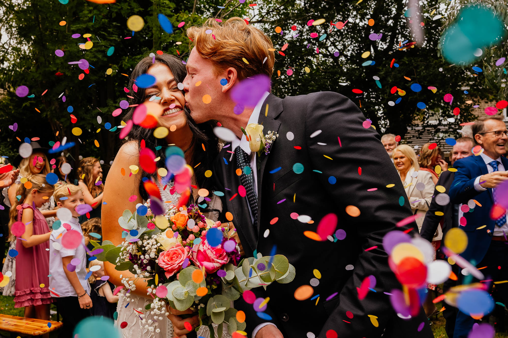 Yorkshire wedding photographer. Groom kissing bride on the cheek with colourful paper confetti in the foreground