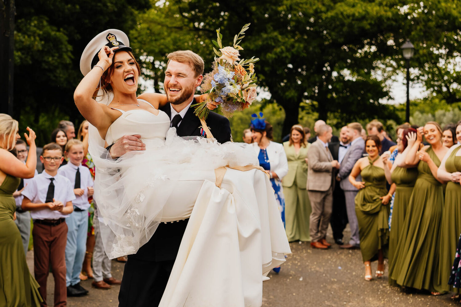 Bride smiling whilst being carried by groom wearing Navy hat
