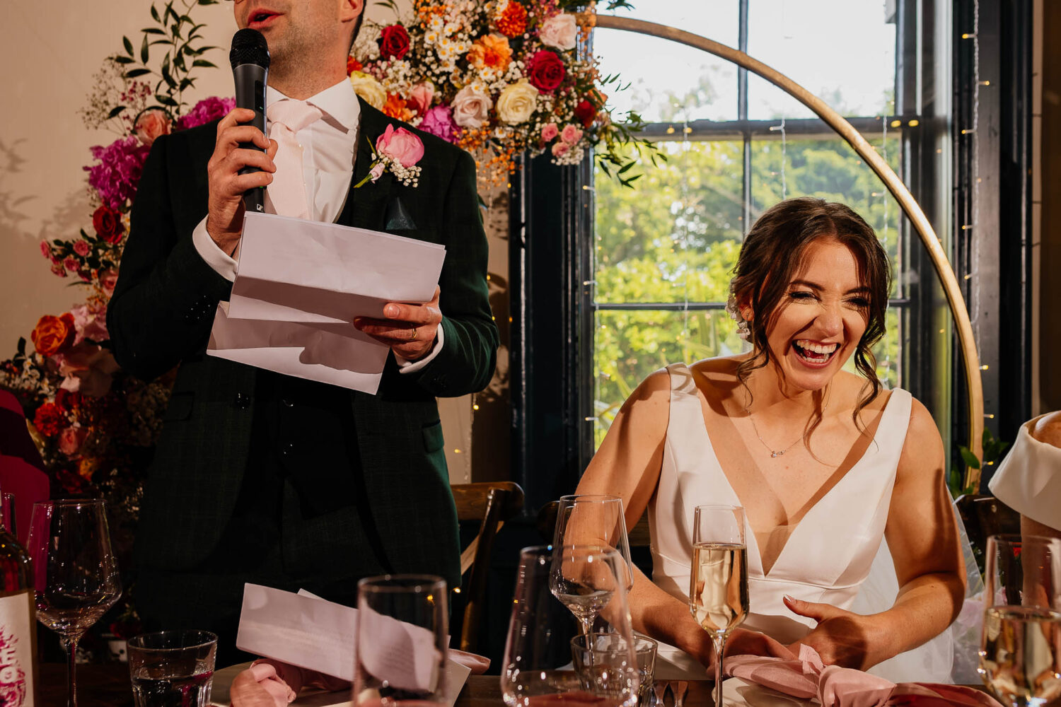 Bride laughing whilst groom gives his speech next to her