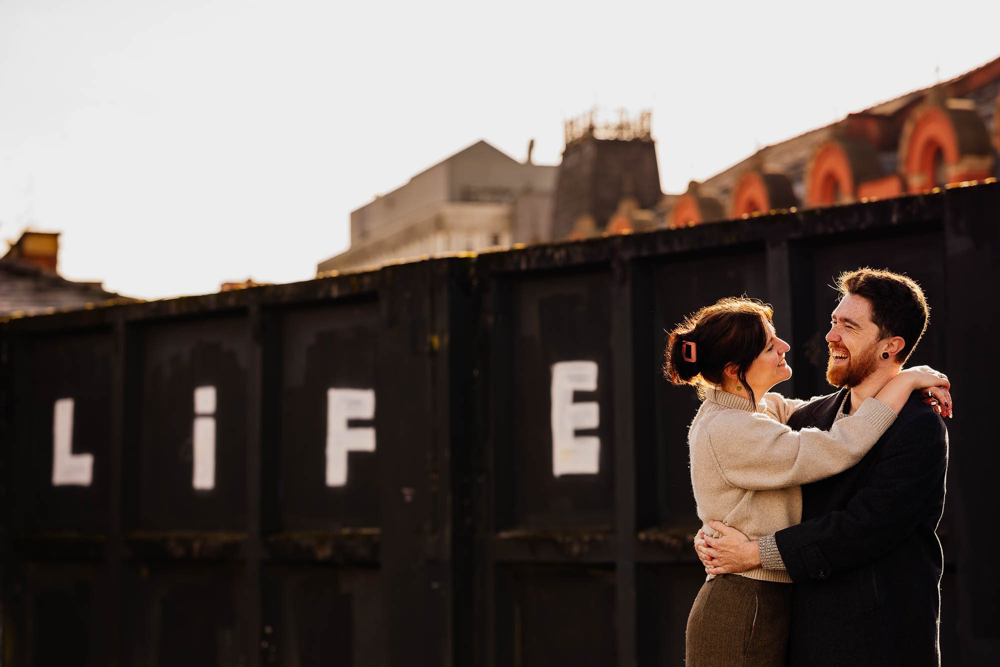 Couple hugging in sun light with 'life' graffiti behind on wall
