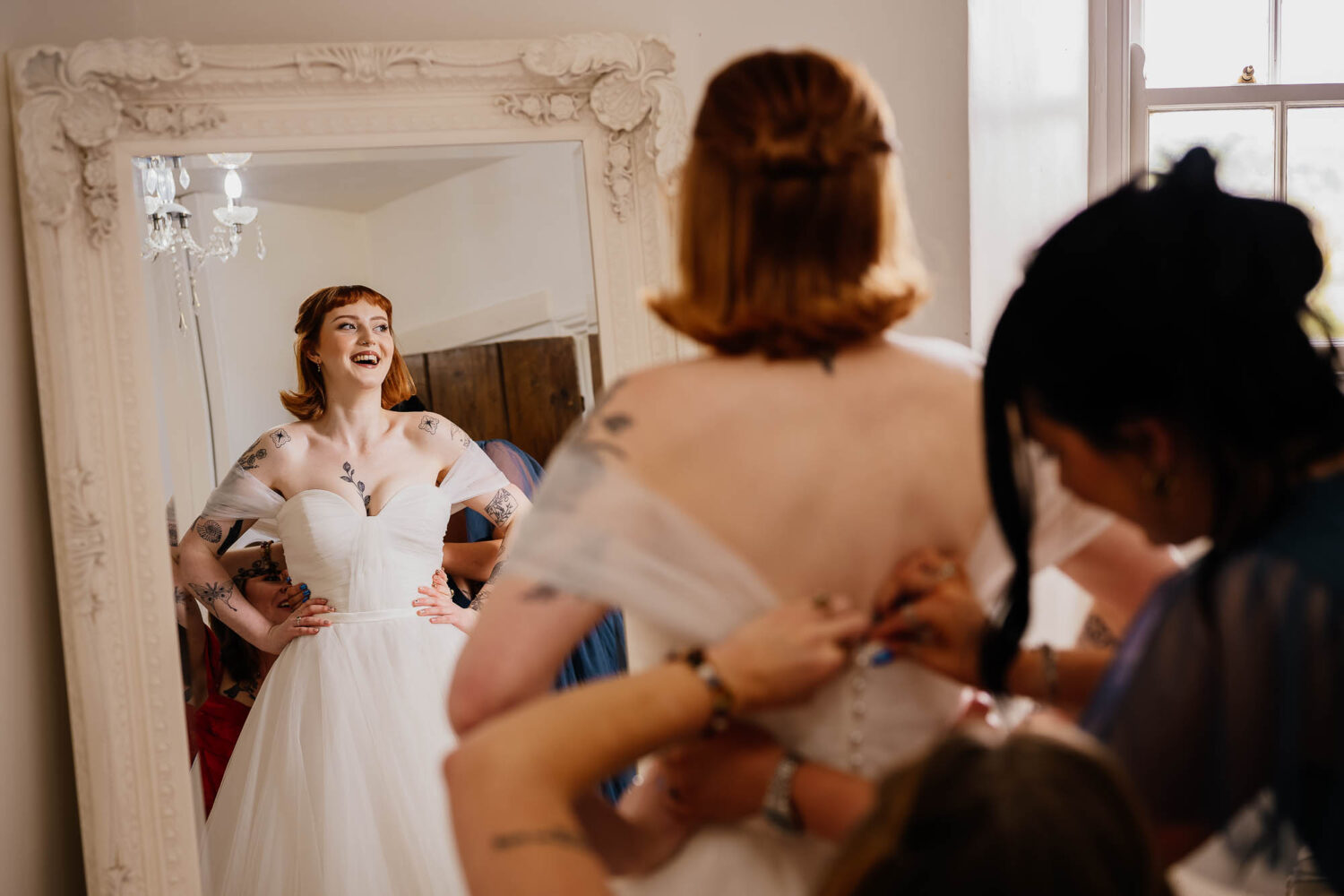 Bride laughing in mirror whilst bridesmaids fasten dress