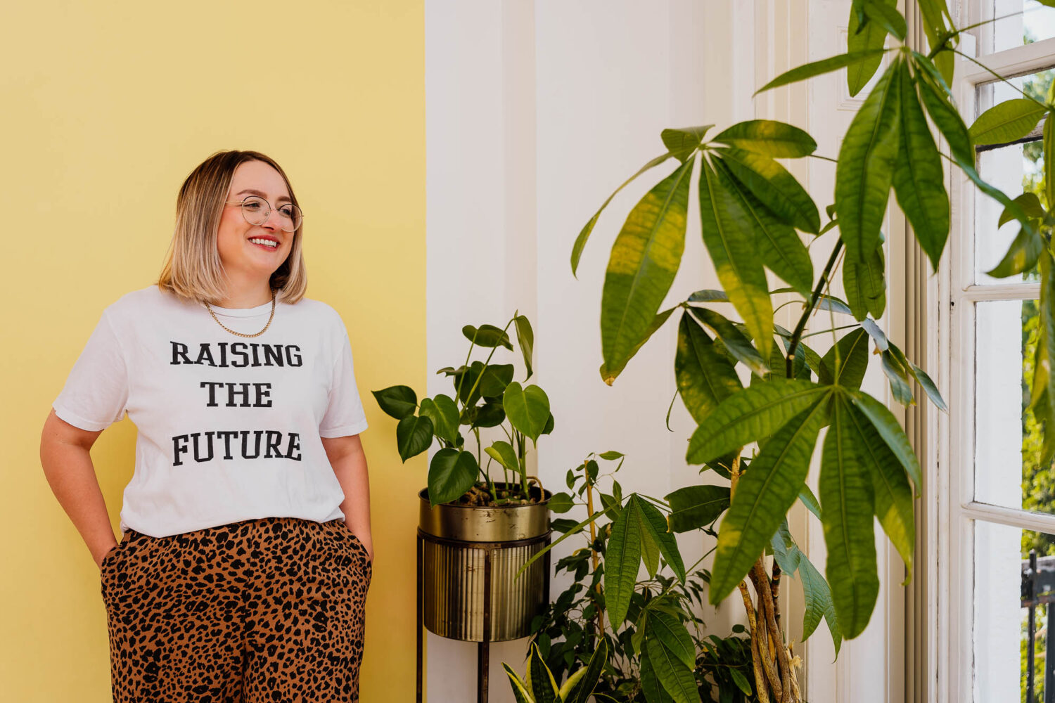 Woman wearing raising the future t-shirt with plants in front of yellow wall