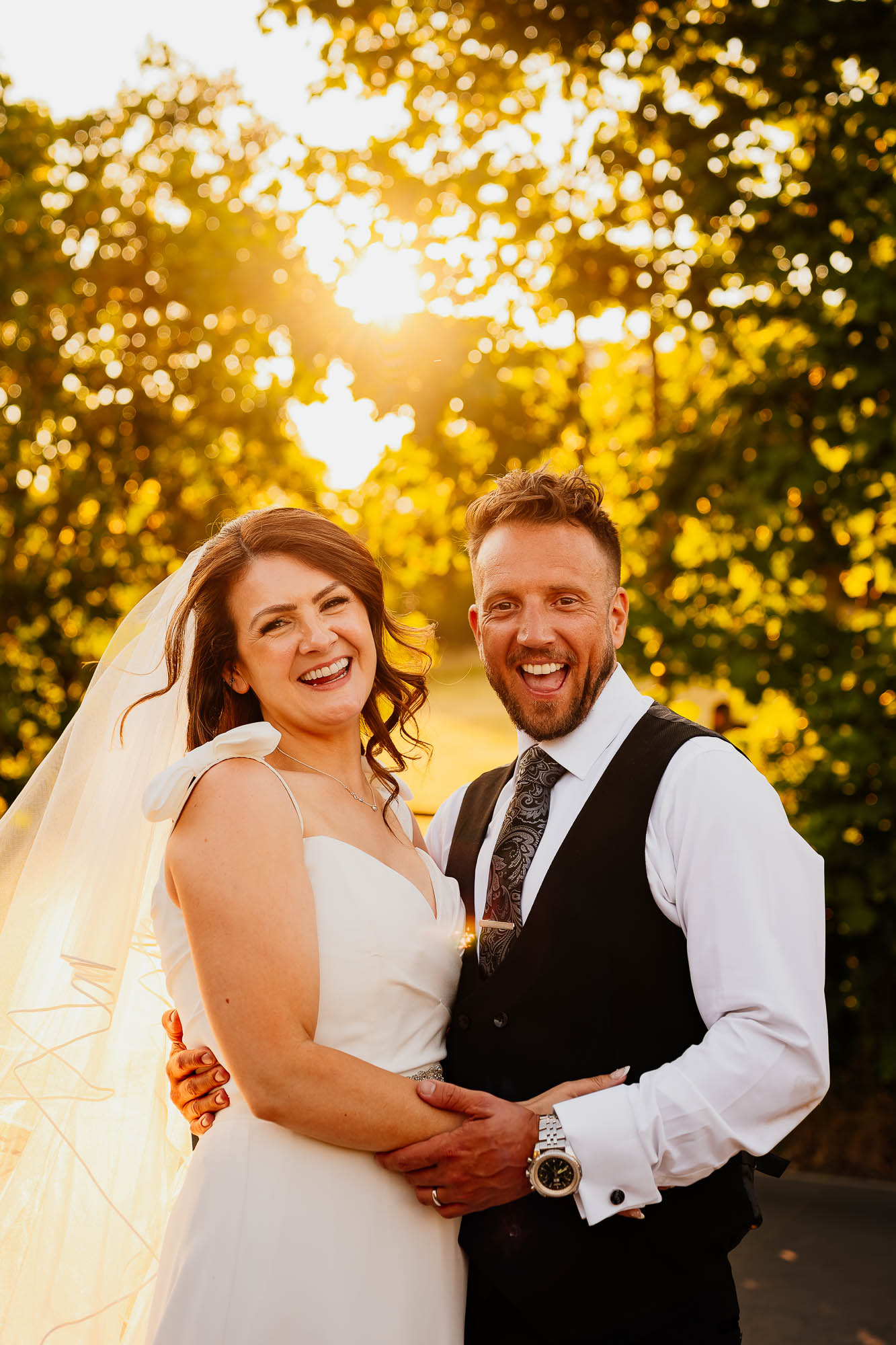 Bride and groom smiling looking at the camera with sun backlighting through the trees