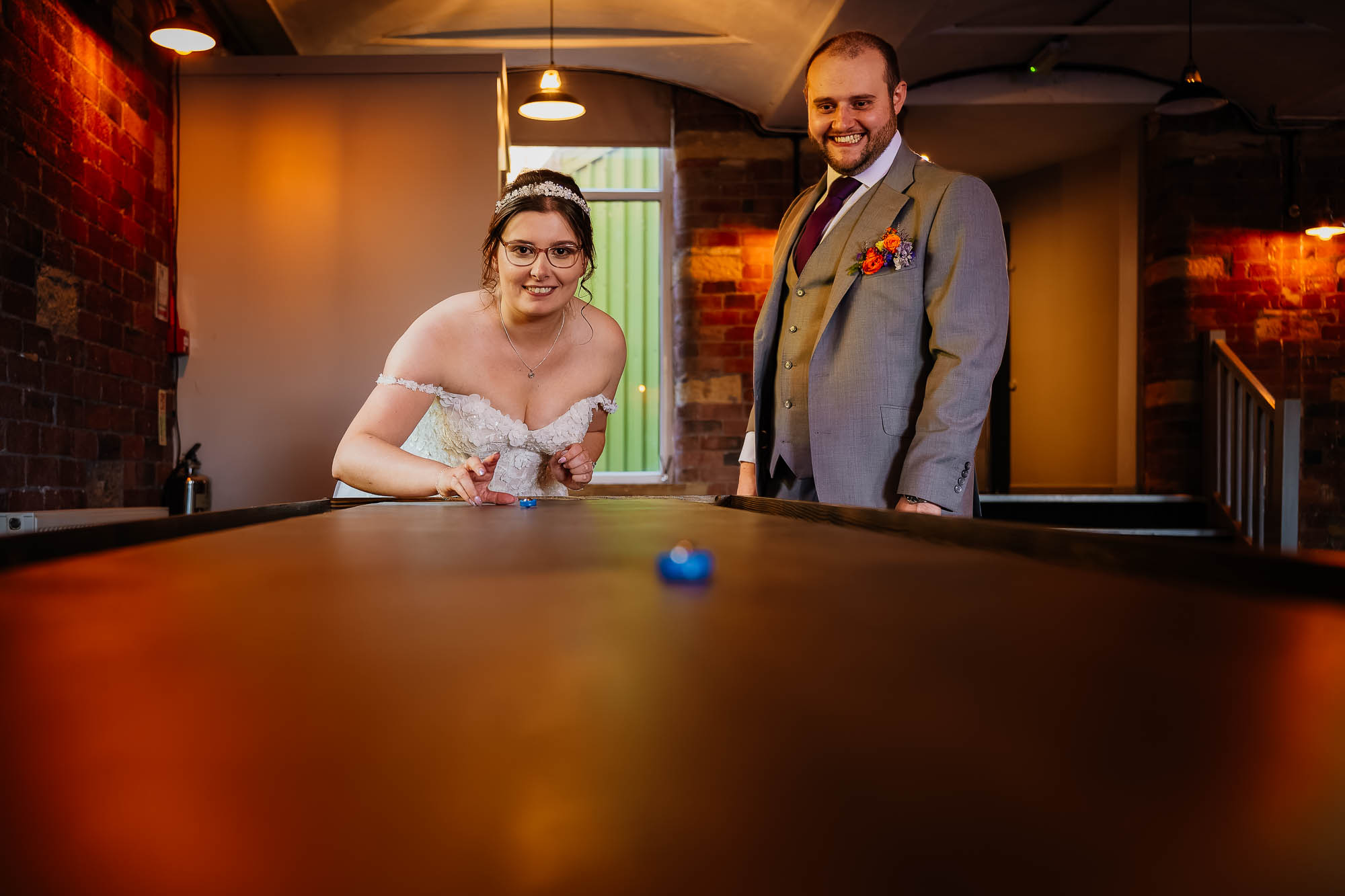 Bride and groom smiling while playing shuffleboard at their indoor wedding reception.