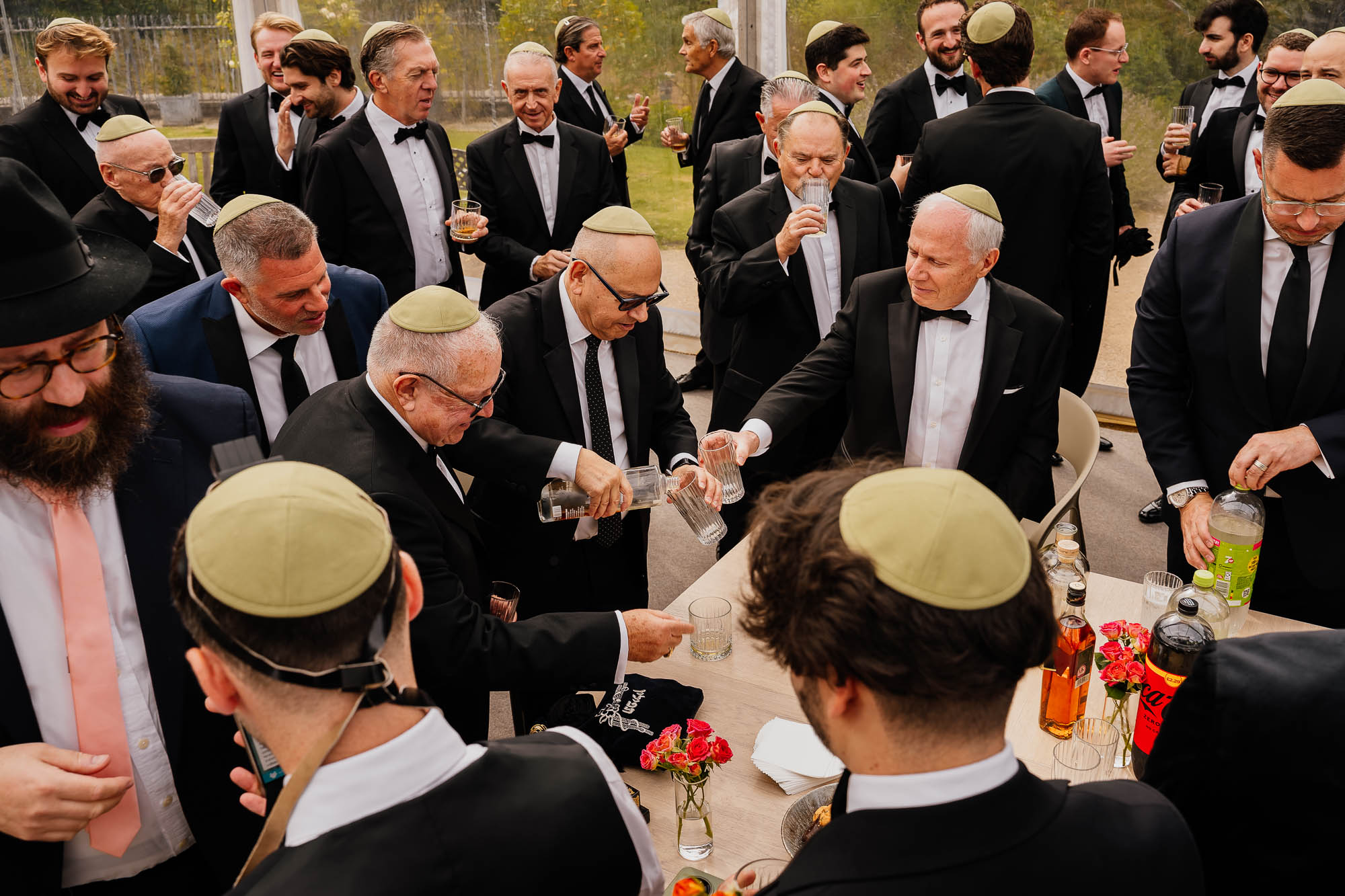 Men in tuxedos wearing kippahs gather around a table pouring drinks during a formal Jewish wedding reception.