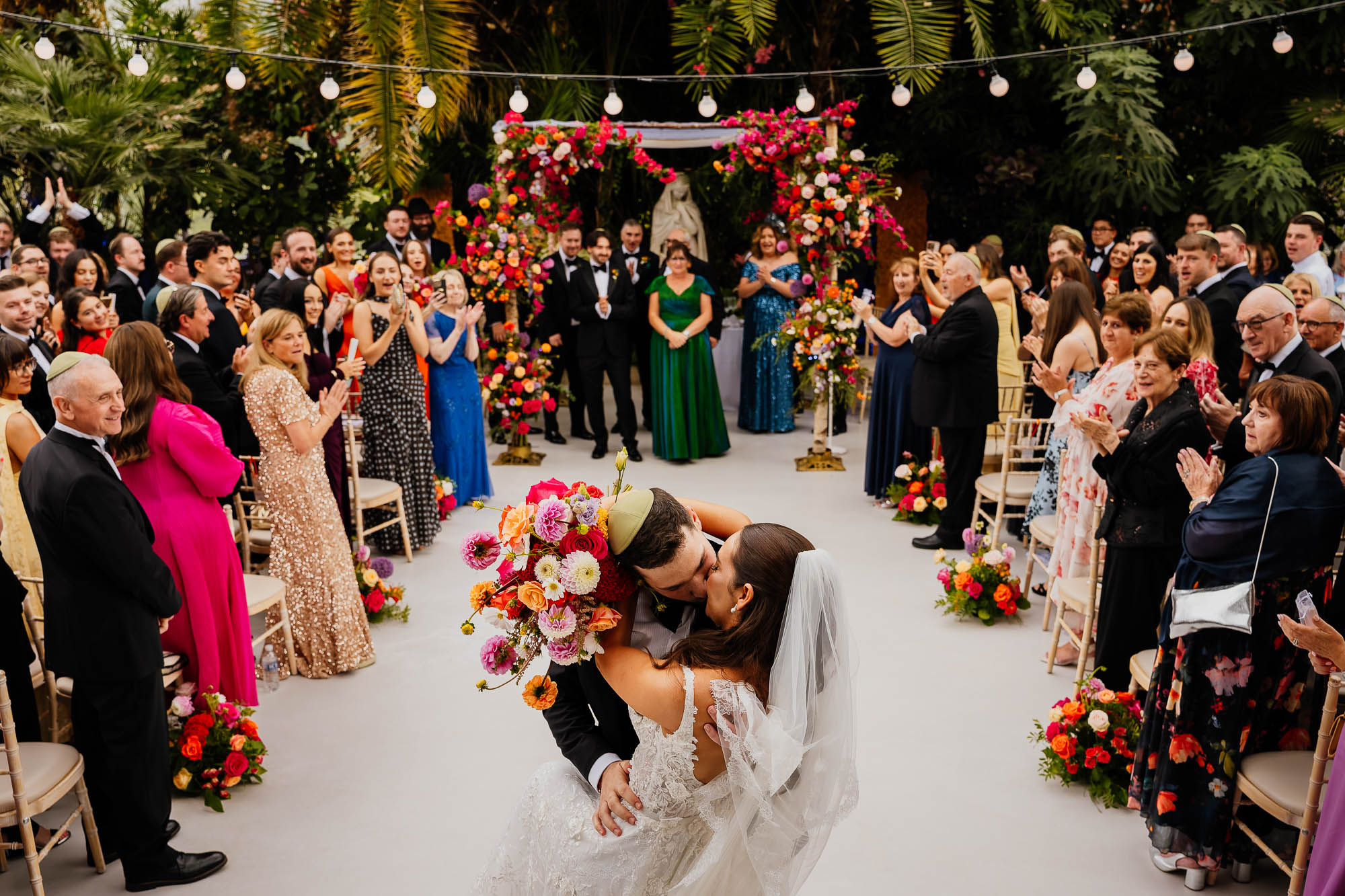 Bride and groom in dip kiss surrounded by guests clapping and bright florals at The Palm House