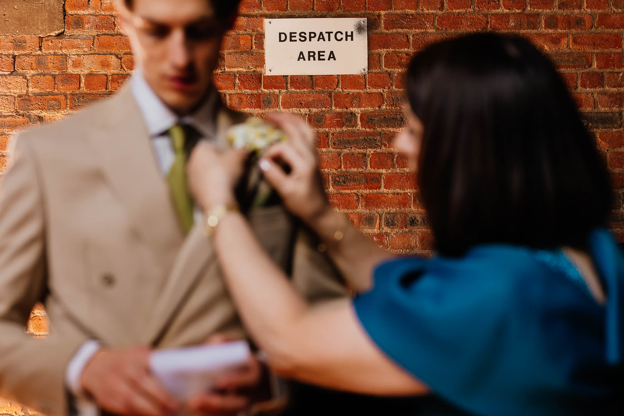 Woman adjusting a boutonniere on a groom's suit jacket in front of a brick wall with a Despatch Area sign.