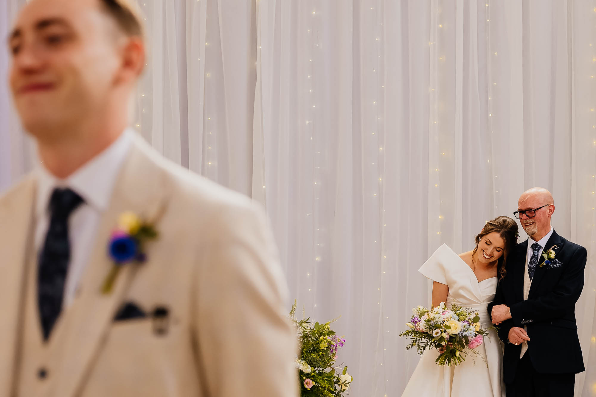 Smiling bride leaning on father's shoulder with bouquet as groom waits in foreground during indoor wedding ceremony.