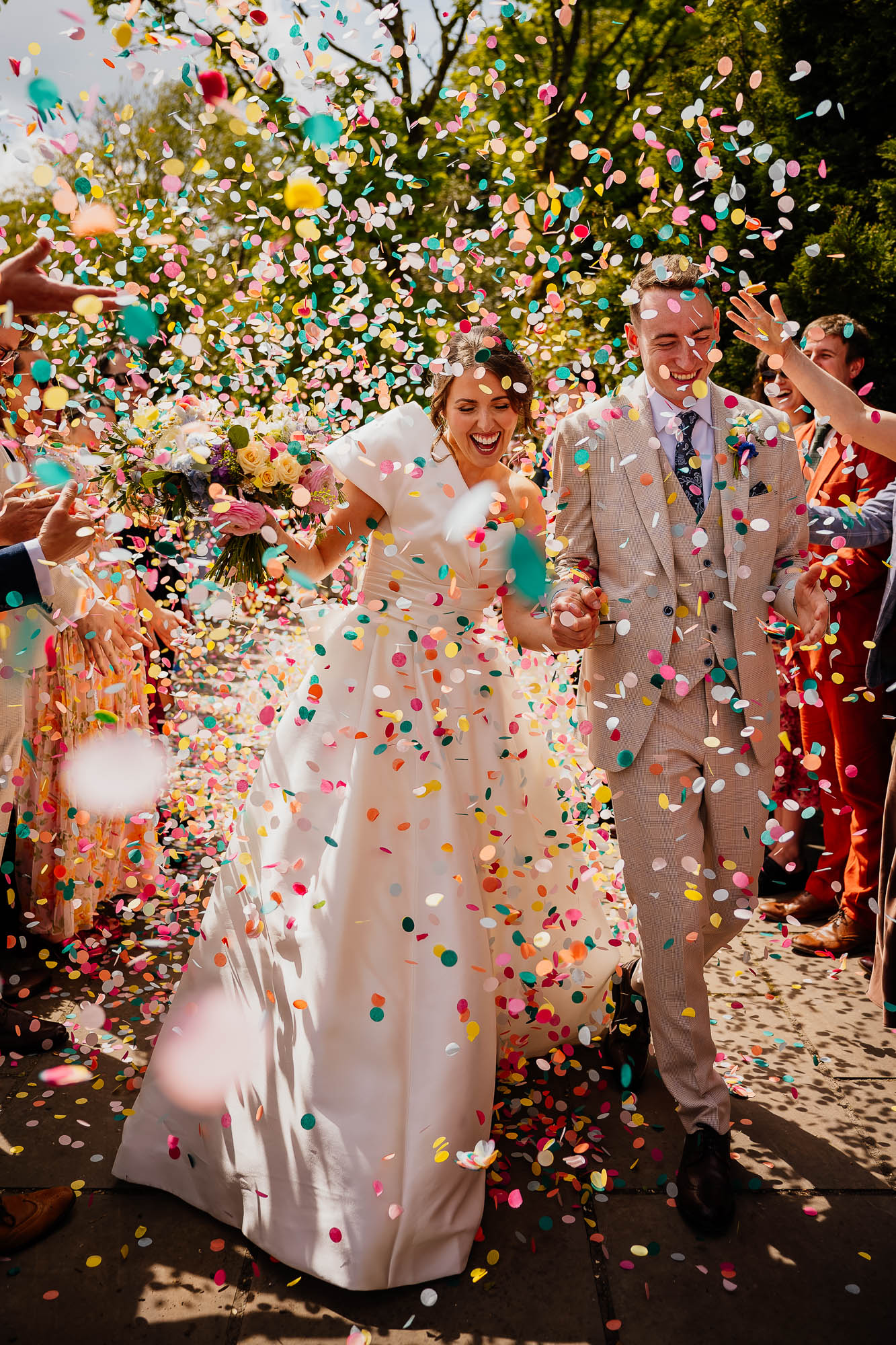 Joyful bride and groom walking through a shower of colorful confetti at an outdoor wedding celebration.
